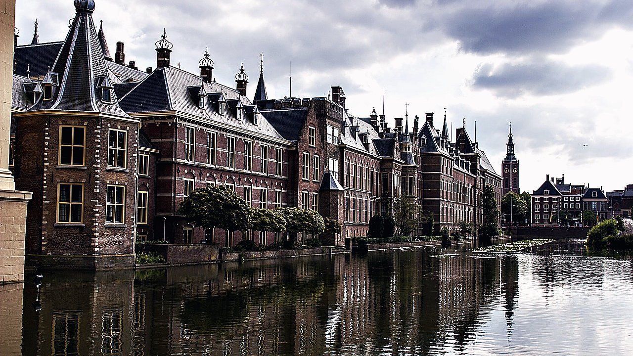 Historische bakstenen gebouwen van het Binnenhof in Den Haag weerspiegelen in de Hofvijver onder een bewolkte lucht