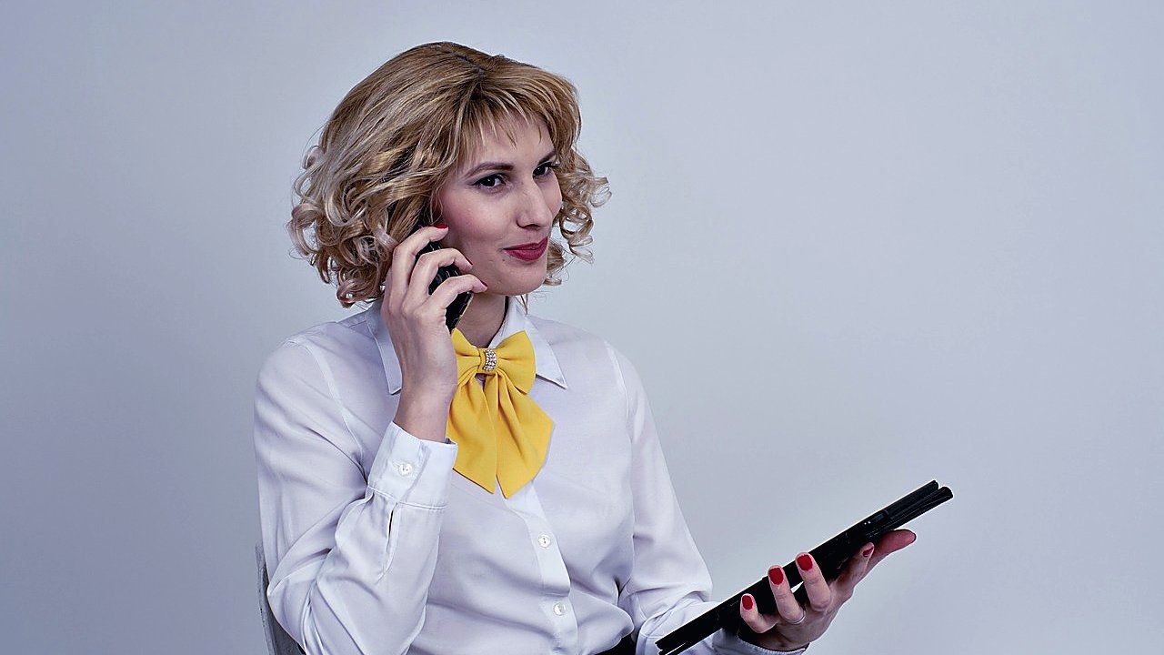 Vrouw met krullend blond haar in wit overhemd en gele strik belt met een telefoon en houdt een tablet vast