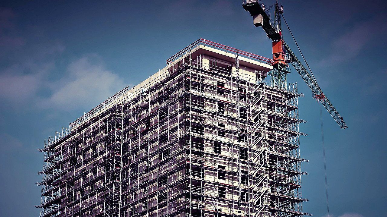 Hoog flatgebouw in aanbouw, volledig omgeven door steigers, met een rood-zwarte torenkraan tegen een blauwe lucht