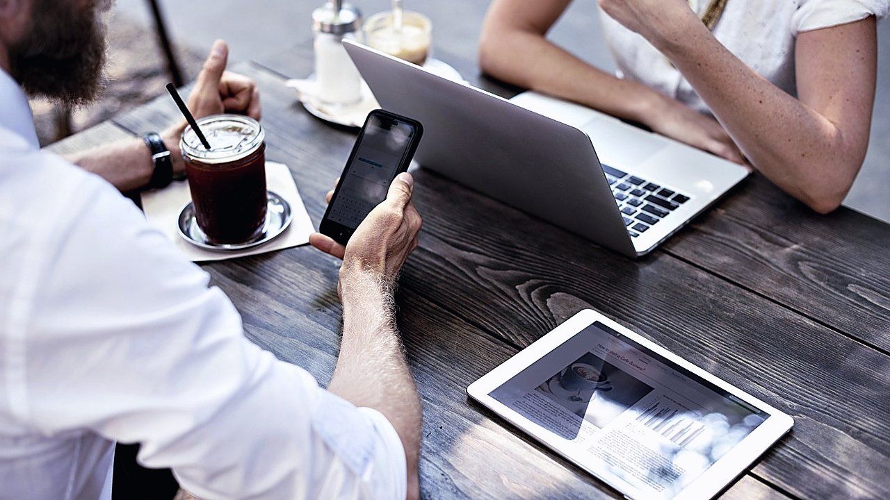 Twee mensen aan een houten caféterras met laptop, tablet, smartphone en een ijskoffie met rietje