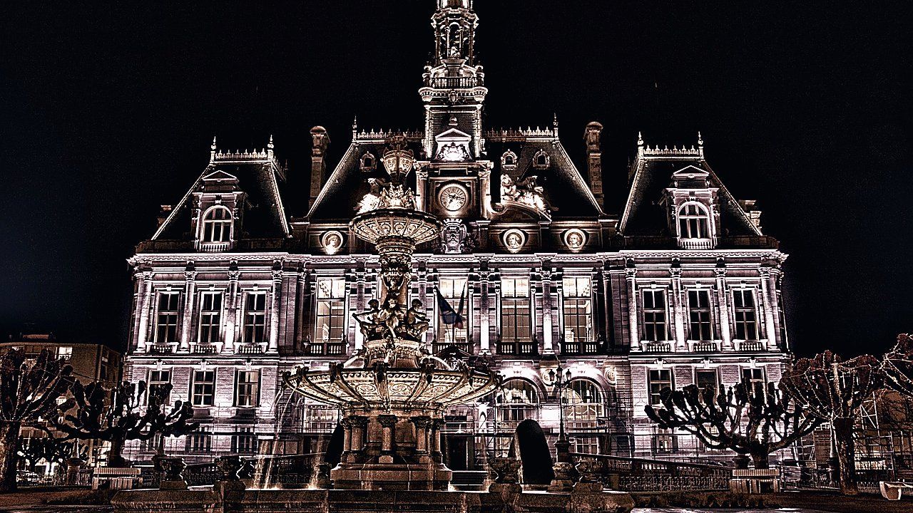 Verlicht stadhuis met klokkentoren in de nacht, met een sierlijke fontein en kale bomen op de voorgrond